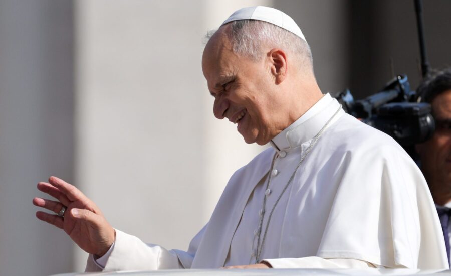 Pápež Lev XIV. v bielom rúchu a s bielou čiapočkou na hlave sa usmieva a zdvíha pravicu v pozdrave, v pozadí je kamera a postava muža.